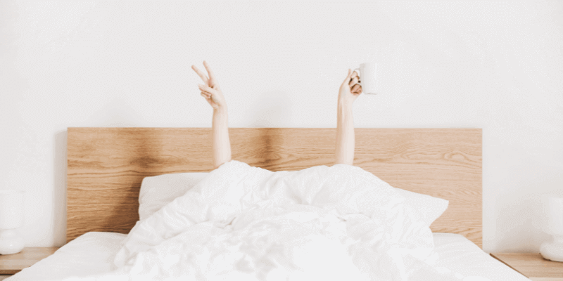 Frau liegt im Bett und streckt die Arme in die Luft, dabei hält sie in der einen Hand eine Kaffeetasse